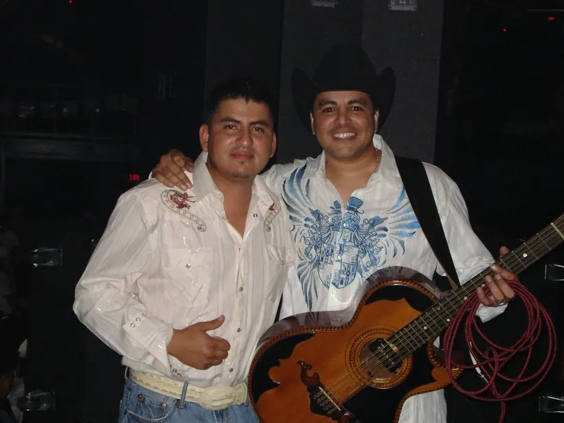 Dos hombres abrazados detrás del escenario, uno con sombrero vaquero negro y una guitarra