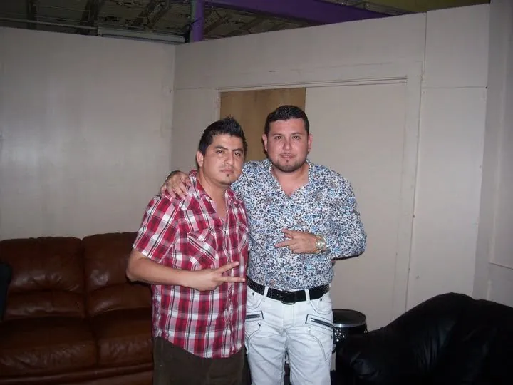 Dos hombres posan frente a un sofá en un camerino, uno con camisa roja a cuadros