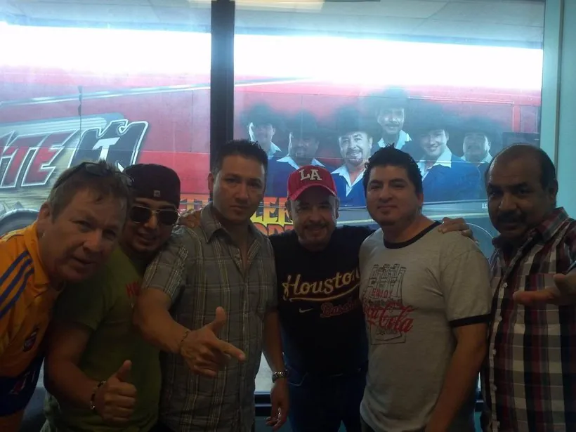 Seis hombres posan en una sala frente a un autobús de gira con el mural de un grupo musical