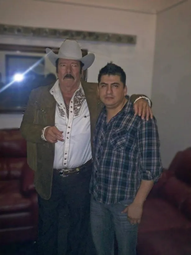 Un hombre con sombrero vaquero blanco abraza a otro con camisa de cuadros dentro de una sala